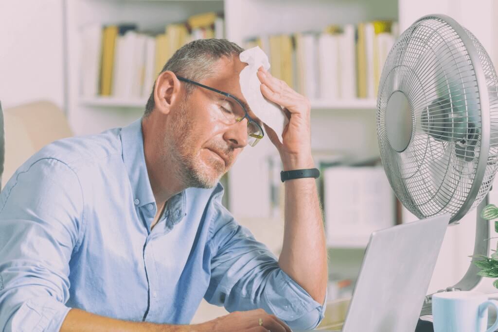 Ein Mann mit hellblauem Hemd sitzt vor einem Ventilator im Büro und wischt sich den Schweiß ab.
