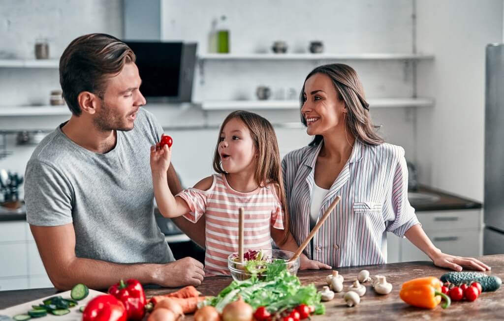 Eine Familie bereitet Gemüse in der Küche zu, ein Kind hält lachend eine Kirschtomate hoch.