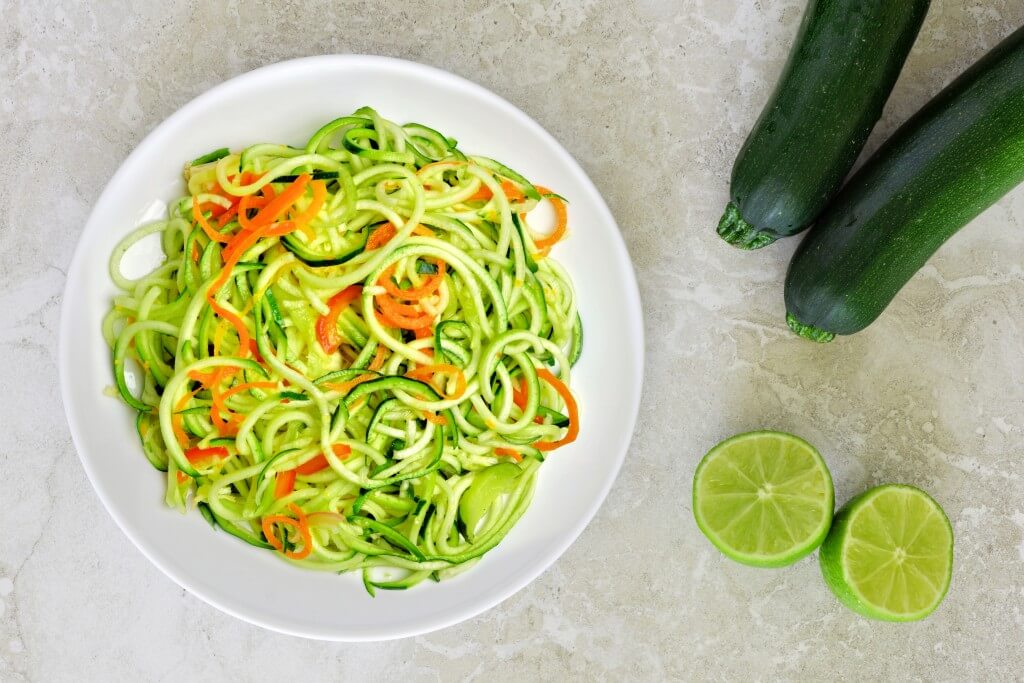 Ein Teller mit Zucchini-Nudeln und Karotten, daneben Zucchini und Limettenscheiben.