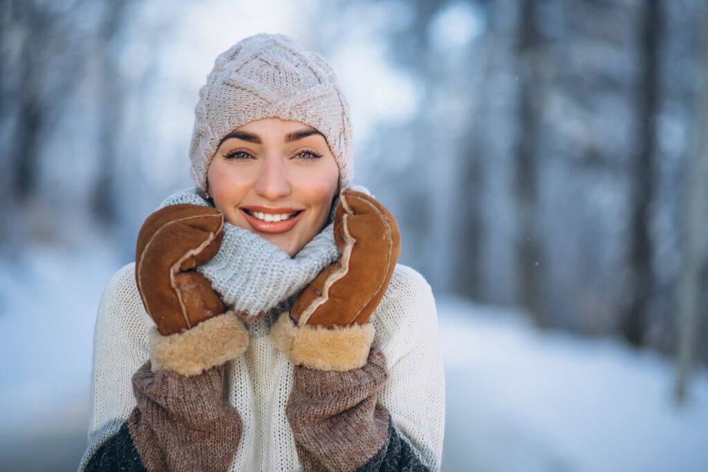 Eine lächelnde Person mit Wintermütze, Schal und Handschuhen im verschneiten Freien.