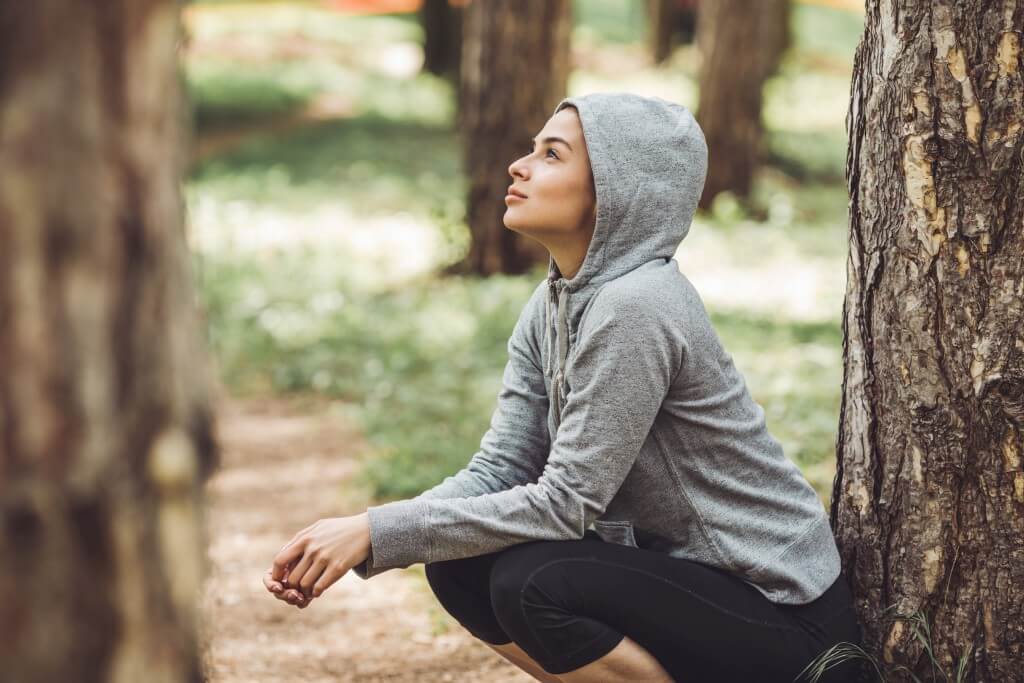 Eine Frau in einem grauen Kapuzenpullover hockt im Wald und blickt nach oben.