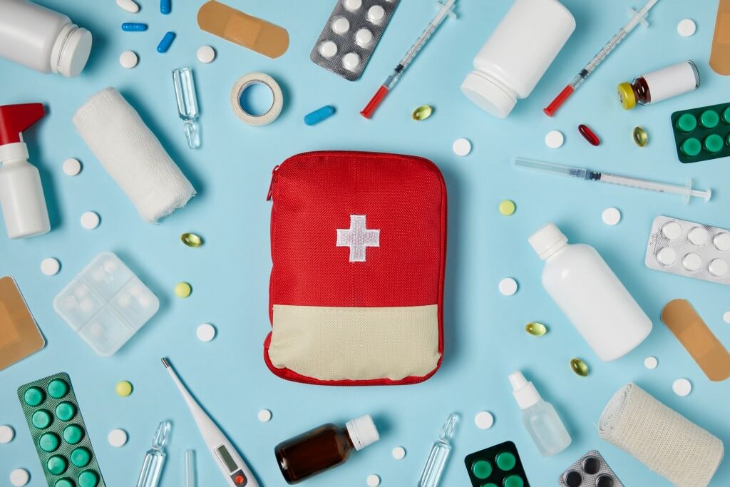 Eine rote Erste-Hilfe-Tasche umgeben von verschiedenen Medikamenten und medizinischen Utensilien.