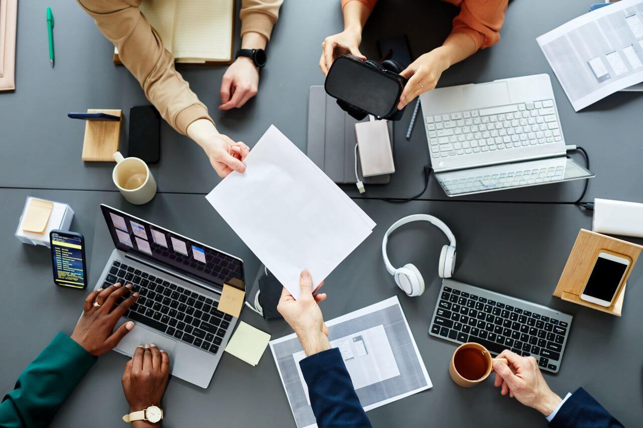 Eine Gruppe von Personen arbeitet mit Laptops und Smartphones an einem gemeinsamen Tisch und tauscht Dokumente aus.