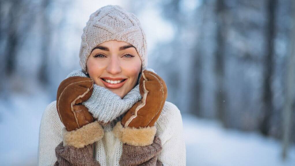 Eine lächelnde Person mit Wintermütze, Schal und Handschuhen im verschneiten Freien.