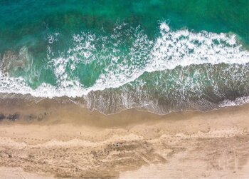 Drohnenansicht einer Meereswelle, die am Übergang vom türkisfarbenen Wasser zum hellbraunen Sandstrand bricht.
