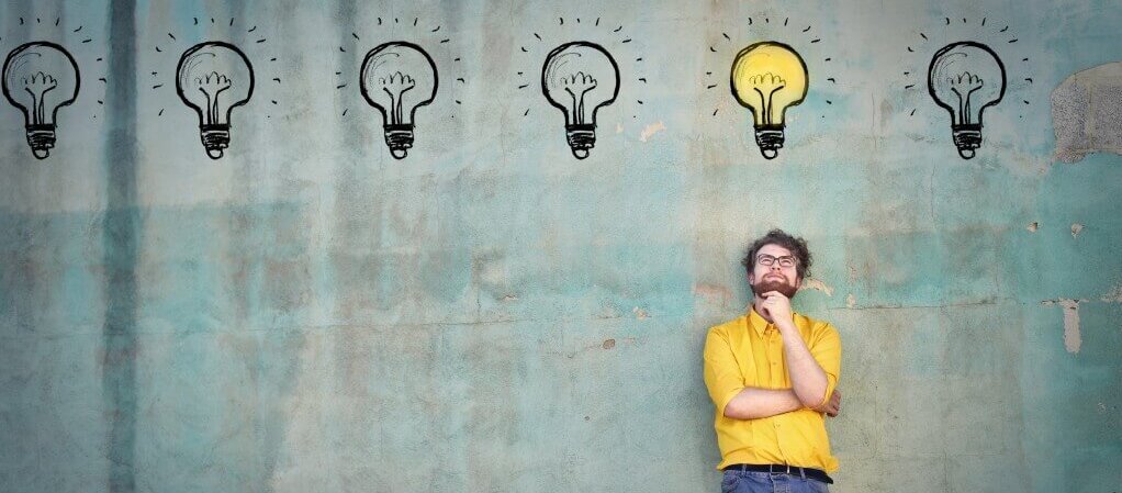 Ein Mann steht vor einer Wand mit Glühbirnen-Zeichnungen; eine davon leuchtet hell, was eine Idee darstellt.