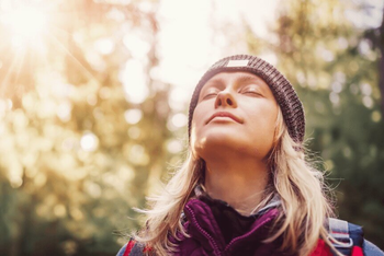  Eine Person mit Mütze und Outdoor-Kleidung steht mit geschlossenen Augen im Sonnenlicht eines Waldes.
