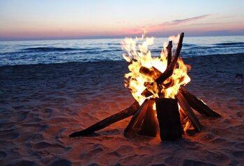 Ein Lagerfeuer brennt am Sandstrand bei Sonnenuntergang, mit ruhigem Meer im Hintergrund.