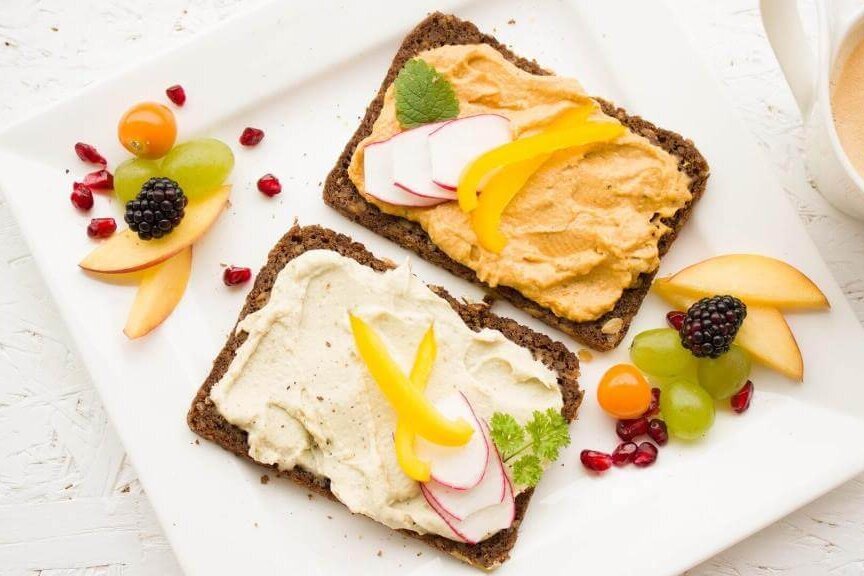 Zwei belegte Brote mit Hummus und Frischkäse, garniert mit Obst, auf einem weißen Teller.
