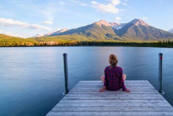 Eine Person sitzt auf einem Holzsteg an einem See und blickt auf ferne Berge unter einem weiten Himmel.