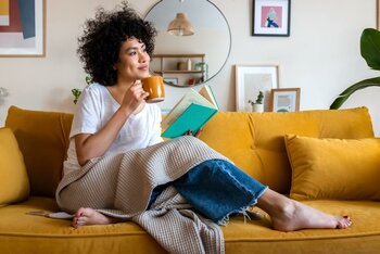 Eine Person sitzt auf einem Sofa, trinkt aus einer Tasse und liest ein Buch.