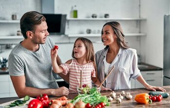Eine Familie bereitet Gemüse in der Küche zu, ein Kind hält lachend eine Kirschtomate hoch.
