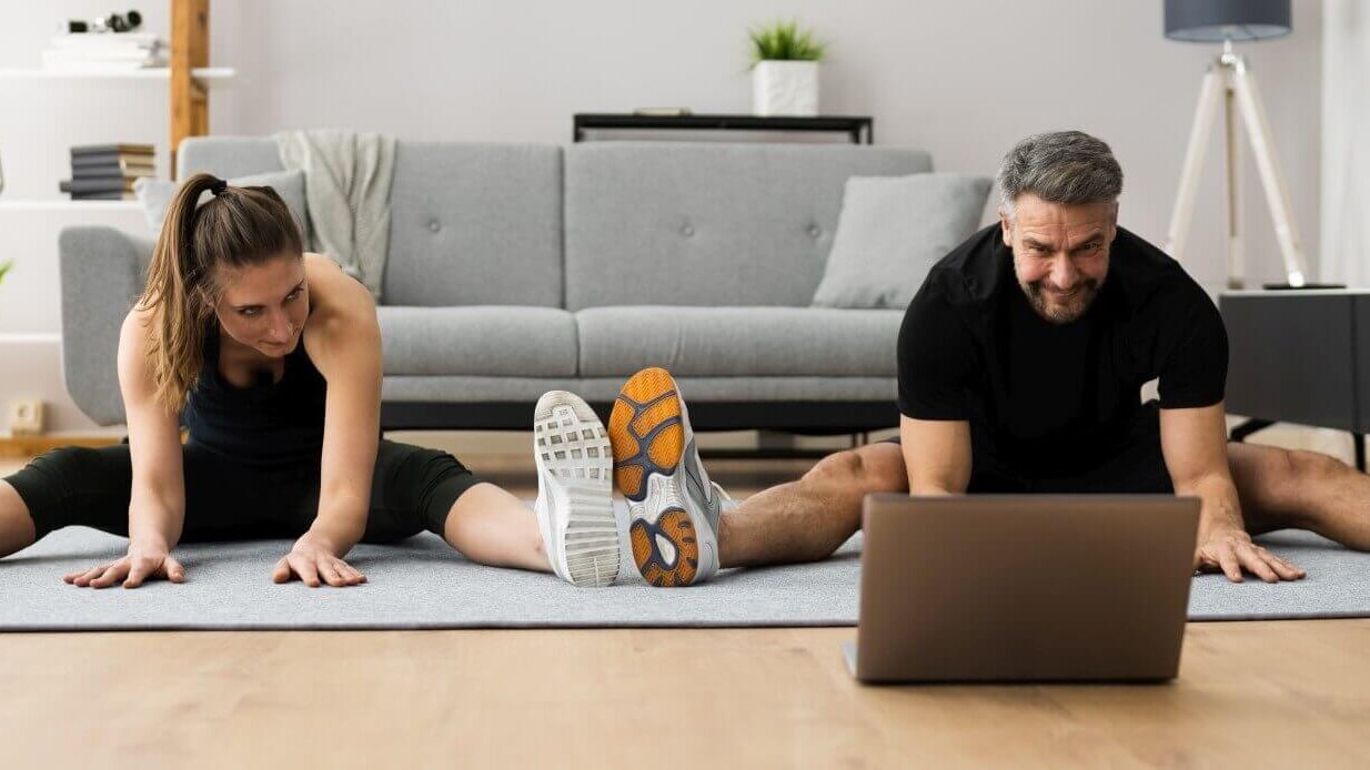Eine Frau und ein Mann machen Dehnübungen auf Matten vor einem Laptop im Wohnzimmer.