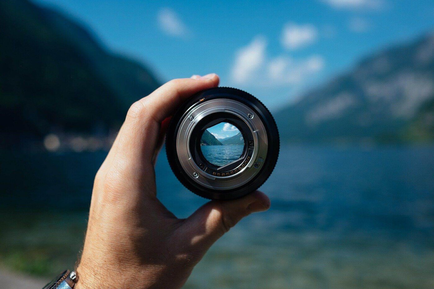 Eine Hand hält ein Kameraobjektiv, das eine klare Sicht auf eine See- und Berglandschaft bietet, Fokus symbolisierend.