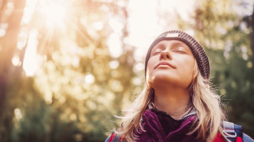  Eine Person mit Mütze und Outdoor-Kleidung steht mit geschlossenen Augen im Sonnenlicht eines Waldes.