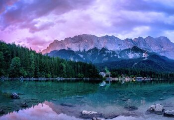 Ein ruhiger See spiegelt einen bewaldeten Berghang und hohe Berge unter einem farbenprächtigen Himmel wider.