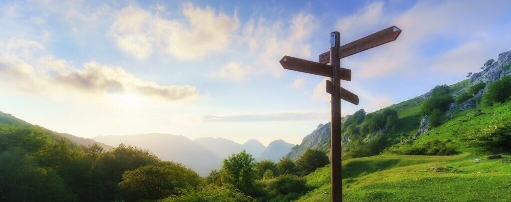 Ein Wegweiser in einer grünen Berglandschaft unter einem hellen Himmel bietet verschiedene Richtungen.