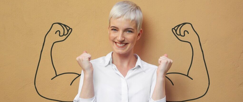 Eine lachende Frau mit kurzen Haaren und gezeichneten Muskelarmen zeigt Stärke und Selbstvertrauen.