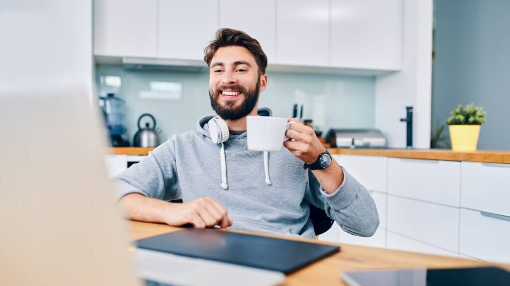 Lächelnder Mann mit Bart und Kopfhörern trinkt Kaffee am Küchentisch.