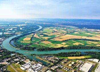 Luftaufnahme einer Flusslandschaft mit weitläufigen Feldern, Wäldern und einem Gewerbegebiet am Ufer.