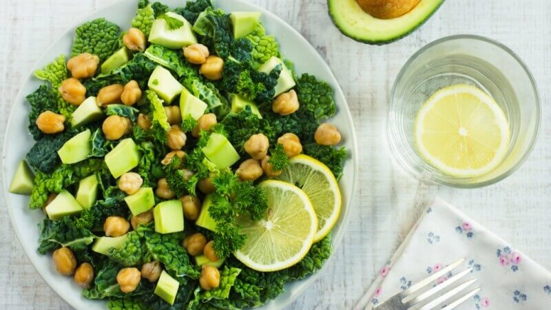 Teller mit Grünkohlsalat, Kichererbsen und Avocado, daneben ein Glas Zitronenwasser.