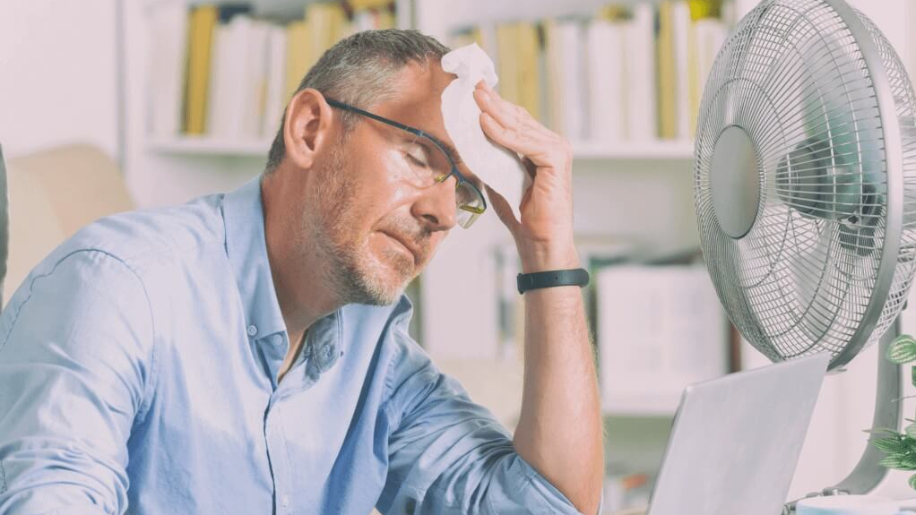 Ein Mann mit hellblauem Hemd sitzt vor einem Ventilator im Büro und wischt sich den Schweiß ab.