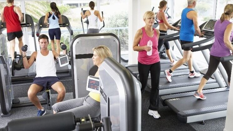 Verschiedene Personen trainieren in einem Fitnessstudio an Laufbändern, Steppern und Kraftgeräten.